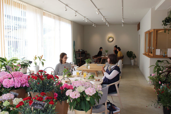 明るいカフェ店内で花に囲まれながら女性客がくつろいでいる風景