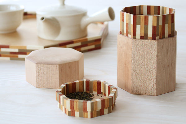 白い急須を背景に、茶葉の入った寄木細工の茶筒と蓋が木製テーブルに並ぶ。