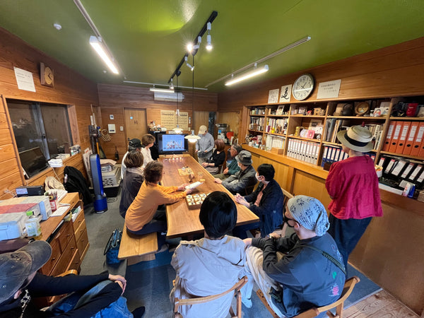 木の質感が感じられる室内で、大勢の人々が長テーブルを囲んで木工について話している様子